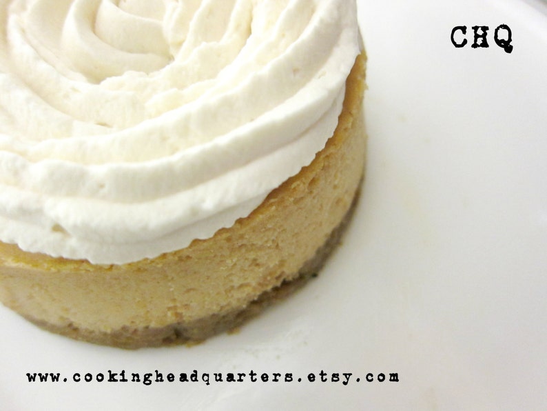 May include: A close-up of a small, round cheesecake topped with a swirl of white whipped cream. The cheesecake is on a white plate with the text "CHQ" and "www.cookingheadquarters.etsy.com" printed on the plate.