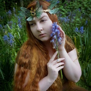 May include: A woman with long red hair wearing a butterfly-themed headpiece, holding a bluebell in a green garden. The headpiece has green butterfly wings and gold accents. The woman is wearing a brown dress, surrounded by bluebells.