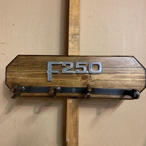 Puede incluir: Un colgador de llaves de madera con un letrero de metal F250 y cuatro ganchos de metal. La madera es de color marrón oscuro y el metal es de color plateado.