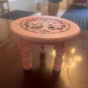 May include: A small pink wooden table with three legs. The table top has a carved tree of life design.