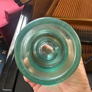 May include: A vintage teal glass electrical insulator featuring a circular, tiered design with a central threaded opening. The object has a smooth, translucent surface with visible internal air bubbles and a thick, rounded outer rim.