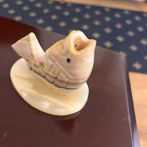 May include: A carved stone fish sculpture with a wide open mouth, resting on an oval base. The fish features tan, brown, and black stripes, and a small black dot for an eye. The base is a light beige color.