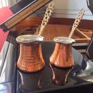 May include: Two copper coffee pots with silver rims and ornate gold-colored handles. The pots have a hammered texture and a classic design. The background includes a piano, suggesting a theme of music and coffee.