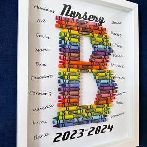 May include: A framed wall art piece featuring the letter B constructed from colorful Crayola crayons. The word Nursery is printed at the top, and the years 2023-2024 are at the bottom. Various names are printed in black ink on both sides of the letter.