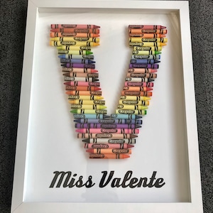 May include: A framed artwork featuring the letter "V" created with colorful crayons. The crayons are arranged in a vertical pattern, forming the shape of the letter. The artwork is signed "Miss Valente" at the bottom.