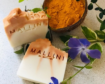 May include: Two bars of natural soap with the word "Mallini" embossed on them. The soap is a light brown color and has a white center. The soap is surrounded by green leaves and a small bowl of yellow turmeric powder.
