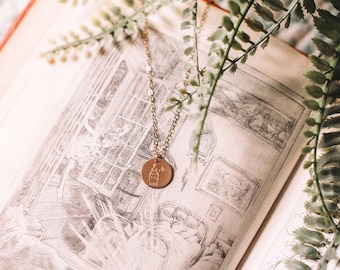 Puede incluir: Un collar de oro con un colgante redondo que presenta una mano con una estrella. El colgante está sobre un libro abierto con una imagen ilustrada.