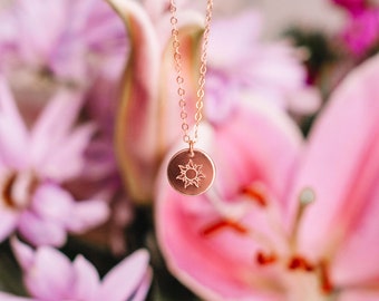 Puede incluir: Un delicado collar de oro con un pequeño colgante redondo que presenta un diseño de sol. El collar está fotografiado sobre un fondo de flores rosas y moradas.
