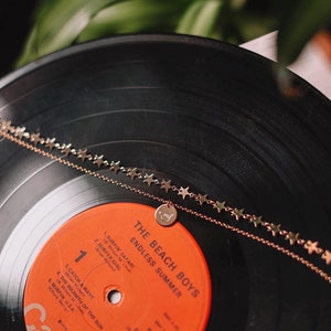 Może przedstawiać: Płyta winylowa z winylu vintage The Beach Boys zatytułowana "Endless Summer". Płyta znajduje się na czarnym gramofonie z złotym naszyjnikiem z gwiazdami i zawieszką z gestem dłoni.