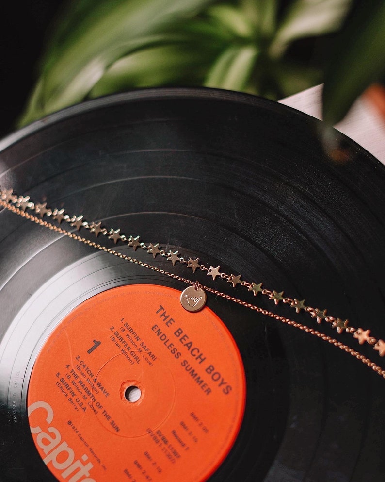 Może przedstawiać: Płyta winylowa z winylu vintage The Beach Boys zatytułowana "Endless Summer". Płyta znajduje się na czarnym gramofonie z złotym naszyjnikiem z gwiazdami i zawieszką z gestem dłoni.