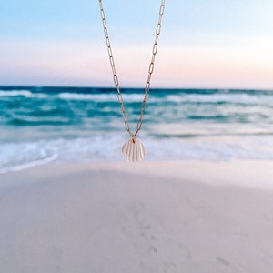 Może przedstawiać: Złoty łańcuszek z białym muszlowym wisiorkiem wiszącym na rozmytym tle plaży i oceanu.