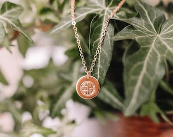 Puede incluir: Un collar dorado con un colgante circular con un diseño grabado. El collar cuelga sobre un fondo de hojas verdes de hiedra y un fondo borroso. La cadena es delicada y el colgante está centrado.