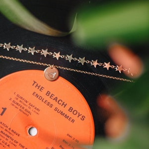 Może przedstawiać: Płyta winylowa z winylu vintage The Beach Boys zatytułowana "Endless Summer". Płyta jest pomarańczowa i czarna z białym tekstem. Złoty naszyjnik z zawieszką w kształcie gwiazdy i małą złotą zawieszką z gestem dłoni leży na płycie.