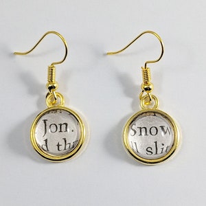 May include: Gold dangle earrings with round glass charms. The charms have a white background with black text. The left earring says "Jon. d th" and the right earring says "Snow ll slit".