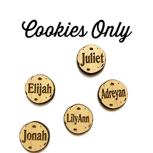 May include: Six wooden cookie-shaped magnets with names written on them. The names are Juliet, Elijah, Adreyan, LilyAnn, and Jonah. The magnets are for a cookie jar and say "Cookies Only" at the top.