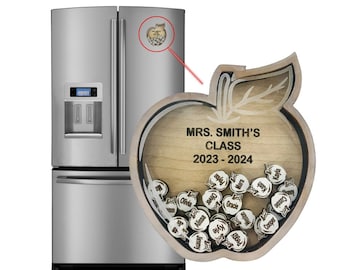 Puede incluir: Caja de madera con forma de manzana con un panel frontal transparente. La caja tiene un recorte de una hoja y un tallo. El panel frontal muestra el texto "MRS. SMITH'S CLASS 2023-2024". Dentro de la caja hay pequeñas piezas de madera con forma de manzana con nombres escritos en ellas.