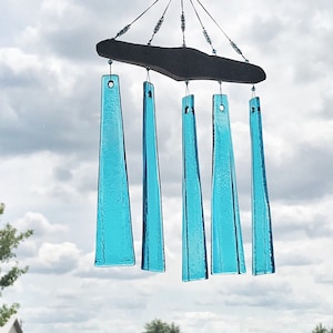 May include: A handcrafted wind chime with five turquoise glass chimes suspended from a dark wooden plank. The chimes are rectangular with a tapered shape, and the sky is visible in the background.