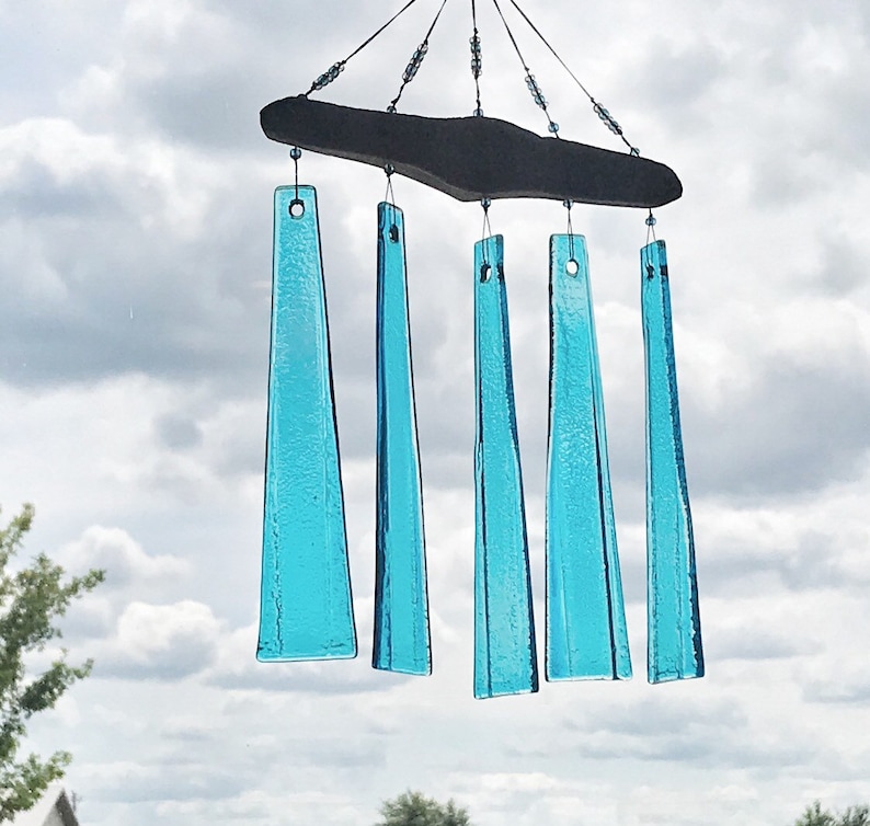 May include: A handcrafted wind chime with five turquoise glass chimes suspended from a dark wooden plank. The chimes are rectangular with a tapered shape, and the sky is visible in the background.