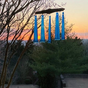 May include: A handmade wind chime with five blue glass tubes suspended from a wooden plank. The wind chime hangs from a tree branch against a backdrop of a colorful sunset. The glass tubes are a vibrant blue color.