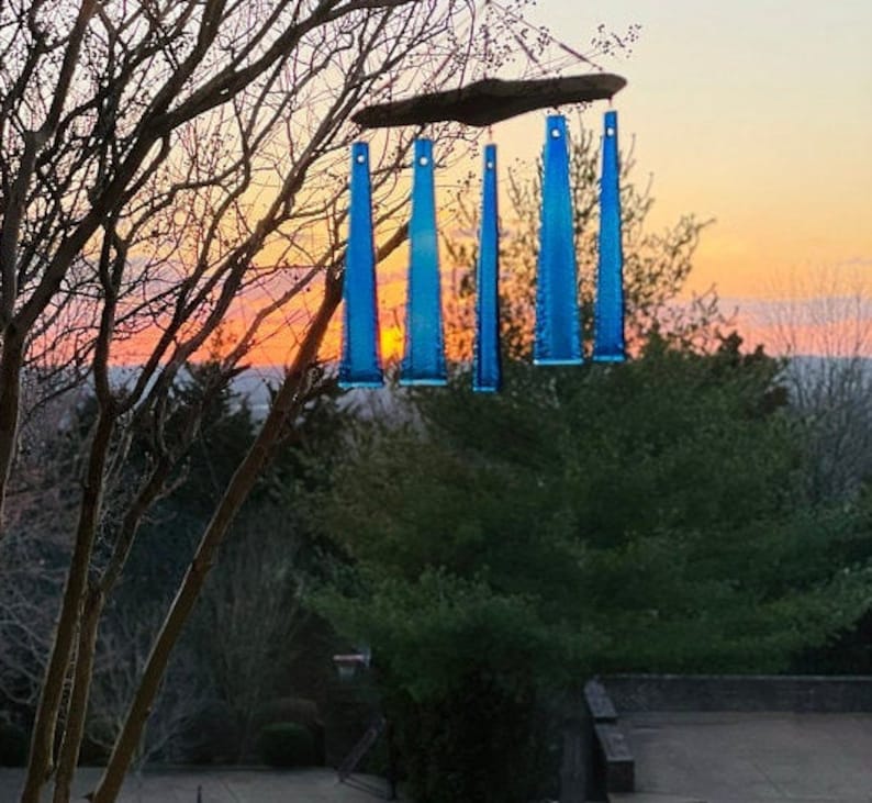 May include: A handmade wind chime with five blue glass tubes suspended from a wooden plank. The wind chime hangs from a tree branch against a backdrop of a colorful sunset. The glass tubes are a vibrant blue color.