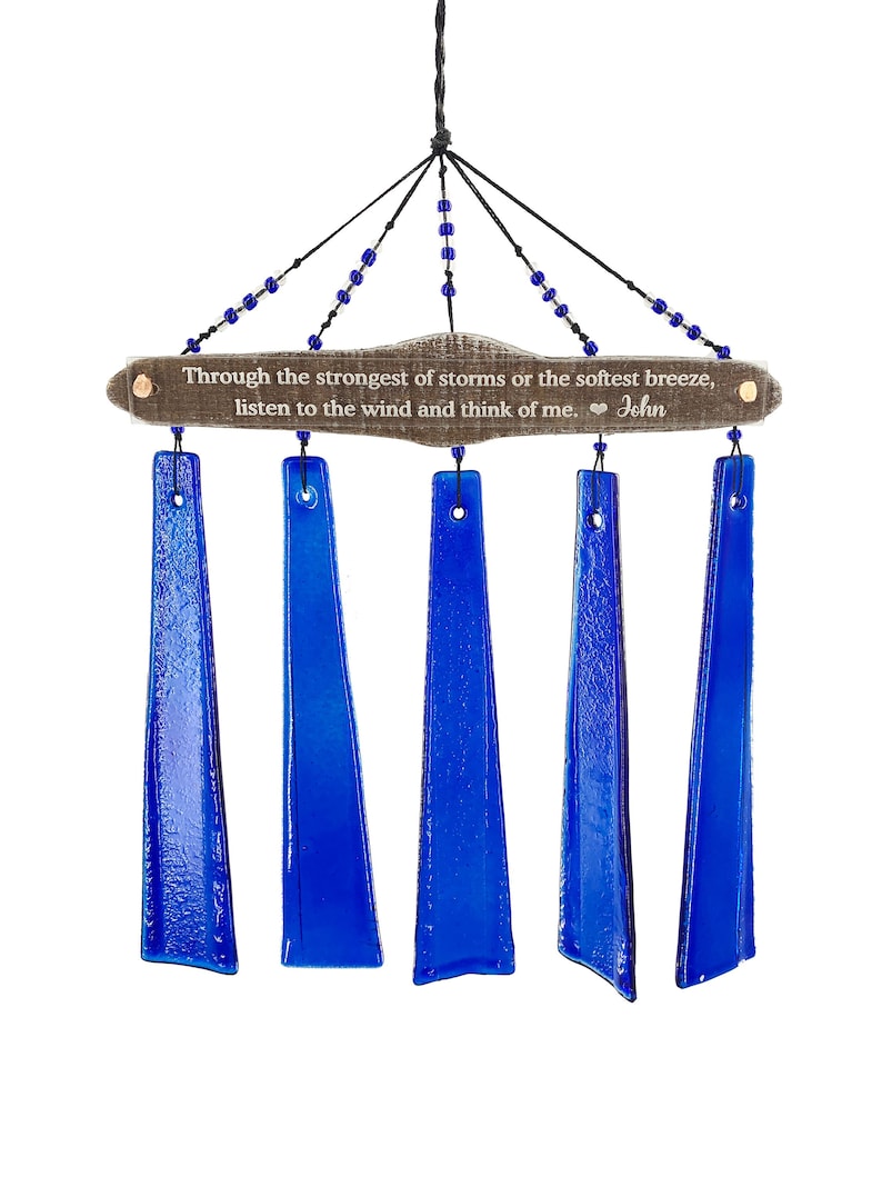 May include: A wind chime with five vibrant blue glass chimes suspended from a wooden plaque. The plaque has the text "Through the strongest of storms or the softest breeze, listen to the wind and think of me. John". Blue beads adorn the hanging strings.