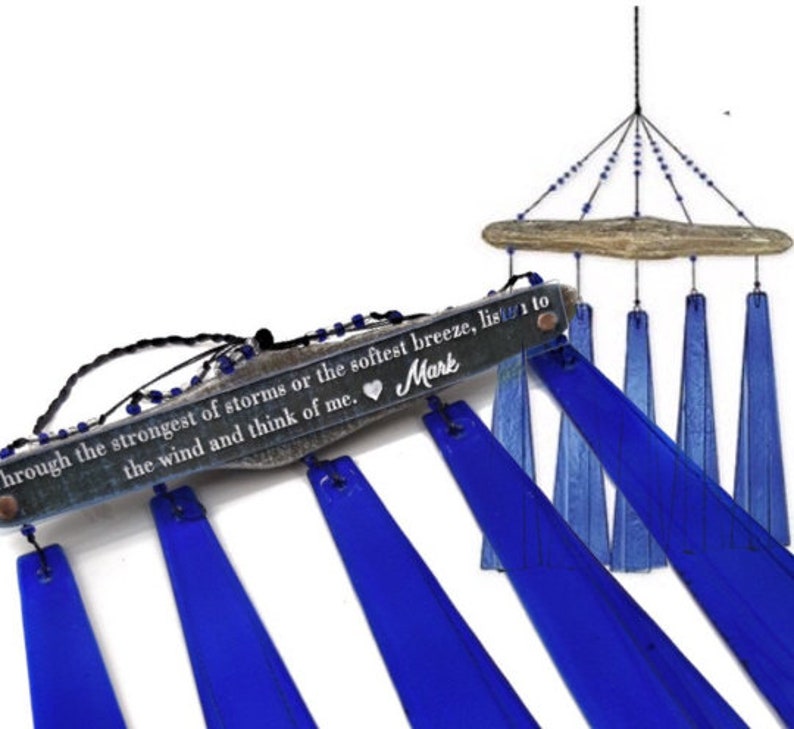 May include: A blue glass wind chime with a wooden top and a decorative inscription. The inscription reads "through the strongest of storms or the softest breeze, listen to the wind and think of me." The wind chime has a heart symbol and the name "Mark".