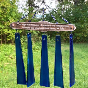 May include: A handcrafted wind chime with five deep blue glass chimes suspended from a wooden plaque. The plaque has the text "Through the strongest of storms or the softest breeze, listen to the wind and think of me. John". Blue and clear beads adorn the top.