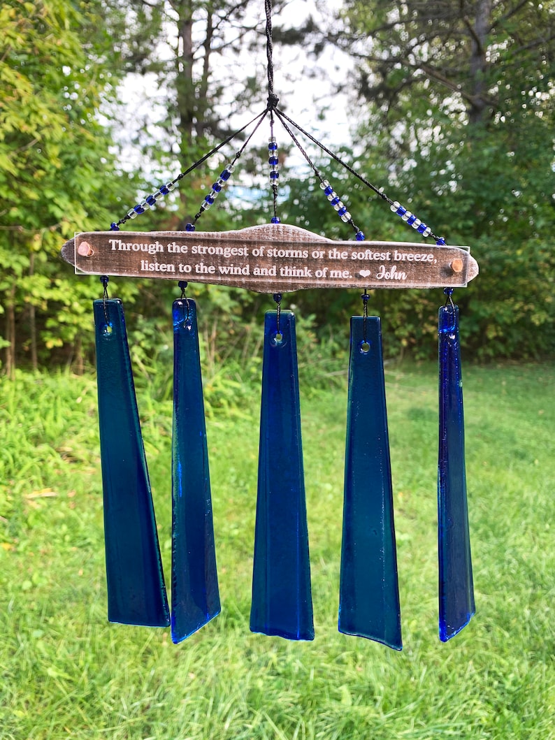 May include: A handcrafted wind chime with five deep blue glass chimes suspended from a wooden plaque. The plaque has the text "Through the strongest of storms or the softest breeze, listen to the wind and think of me. John". Blue and clear beads adorn the top.