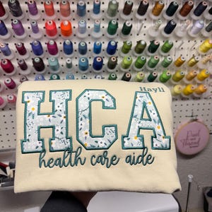 Puede incluir: Sudadera crema con texto bordado y diseño floral. La sudadera presenta las letras "HCA" en un contorno verde azulado con un patrón floral, y las palabras "health care aide" debajo. El nombre "Hayli" está bordado encima de la "A".