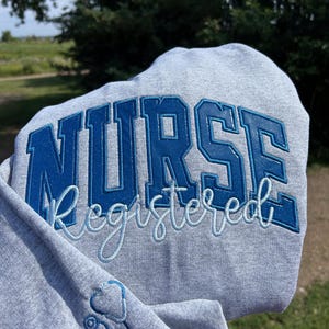 May include: Grey hooded sweatshirt with the text "NURSE registered" embroidered in blue and white. A small blue stethoscope is embroidered on the left sleeve.