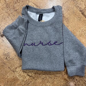 Könnte beinhalten: Ein graues Sweatshirt mit dem Wort "nurse" in lila Schrift auf der Brust gestickt. Das Rundhals-Sweatshirt ist gefaltet, wobei sich die Ärmel überlappen. Das Kleidungsstück ist aus weichem Material.