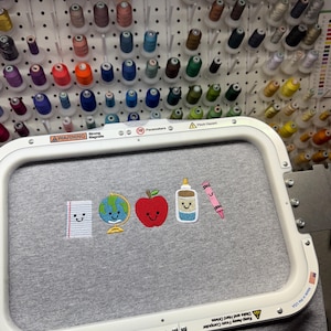 May include: Gray t-shirt with embroidered school-themed patches: notebook, globe, red apple, glue bottle, and pink crayon. The shirt is in a white embroidery hoop. Spools of thread in various colors are in the background.