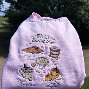 Peut inclure: Sweat-shirt rose clair avec des broderies sur le thème de l'automne. Le motif comprend des feuilles, des pommes, une tarte, des citrouilles et du cidre de pomme, avec le texte "FALL Bucket List".