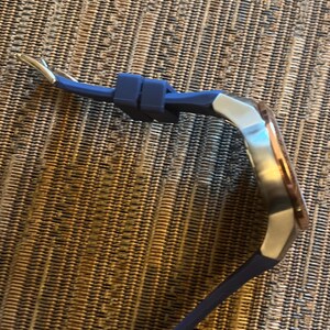 Pu&ograve; includere: Un orologio da polso con cinturino in silicone blu e quadrante argentato e oro rosa. Il cinturino dell'orologio &egrave; fissato con una chiusura argentata. L'orologio &egrave; esposto su una superficie marrone strutturata.