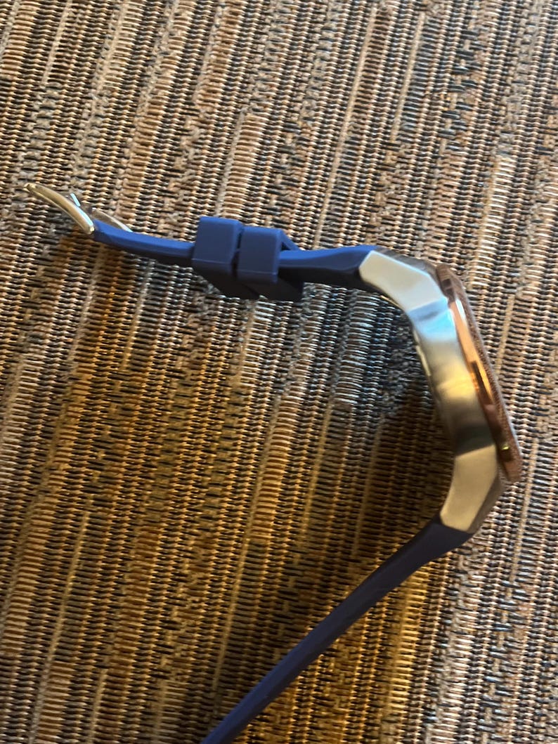 Pu&ograve; includere: Un orologio da polso con cinturino in silicone blu e quadrante argentato e oro rosa. Il cinturino dell'orologio &egrave; fissato con una chiusura argentata. L'orologio &egrave; esposto su una superficie marrone strutturata.