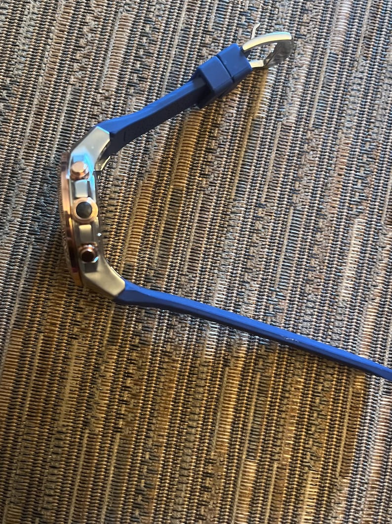 Pu&ograve; includere: Un orologio da polso con cinturino in silicone blu e quadrante argento e oro rosa. L'orologio ha tre pulsanti laterali e una chiusura a fibbia. L'orologio &egrave; appoggiato su una superficie strutturata.