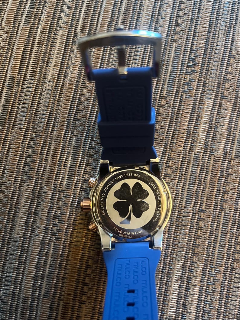 Pu&ograve; includere: Un orologio Mulco in acciaio inossidabile con cinturino in silicone blu. Il quadrante dell'orologio presenta un design a quadrifoglio. L'orologio ha dettagli in oro rosa ed &egrave; visibile il testo "MULCO M10 FOREST MW5-5673-043".