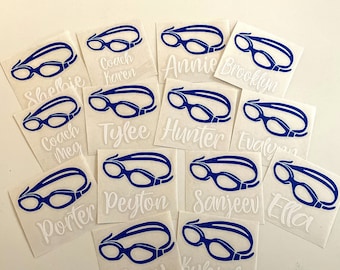 May include: A set of 13 blue and white swim goggles with names written on them. The names include:  'Coach Karen', 'Annie', 'Brooklyn', 'Coach Meg', 'Tylee', 'Hunter', 'Evelyn', 'Porter', 'Peyton', 'Sanjeev', 'Ella', 'Zoey', and 'Kyleigh'.