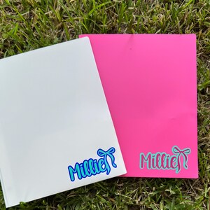 May include: Two rectangular sheets, one white and one bright pink, rest on green grass. Each sheet features the name "Millie" in a cursive font with a bow graphic. The white sheet has blue accents, while the pink sheet has teal accents.