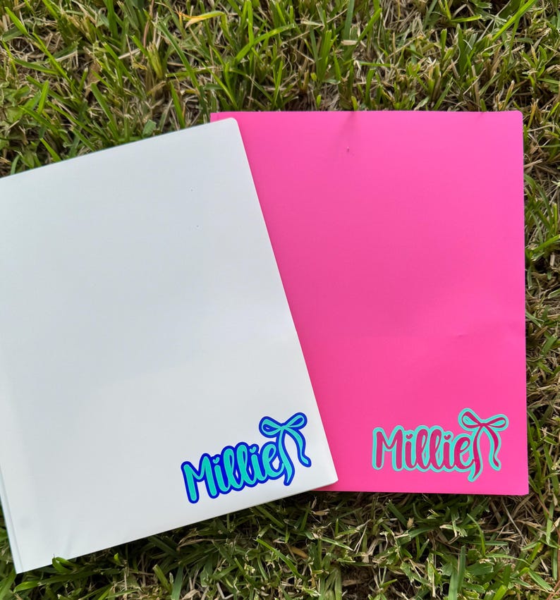 May include: Two rectangular sheets, one white and one bright pink, rest on green grass. Each sheet features the name "Millie" in a cursive font with a bow graphic. The white sheet has blue accents, while the pink sheet has teal accents.
