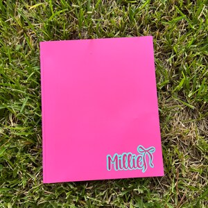May include: A bright pink notebook with the name "Millie" in a teal script font with a bow design. The notebook is rectangular and lies on a bed of green grass.