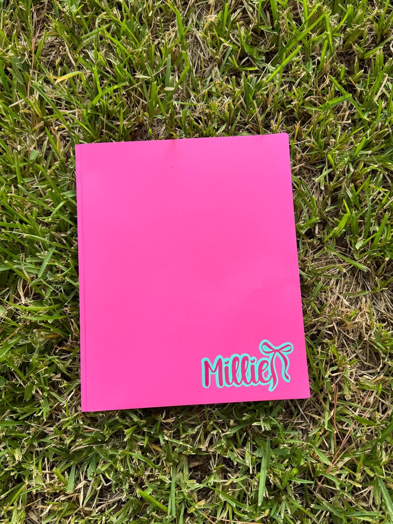 May include: A bright pink notebook with the name "Millie" in a teal script font with a bow design. The notebook is rectangular and lies on a bed of green grass.