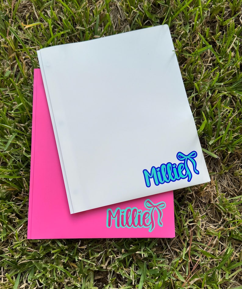 May include: Two notebooks, one white and one pink, are stacked on green grass. The white notebook has a blue and teal sticker that says "Millie" with a bow. The pink notebook has a teal and pink sticker that says "Millie" with a bow.