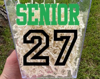 May include: A clear plastic container with a scalloped top edge, filled with light beige shredded paper filler. The front of the container features the word SENIOR in green block letters above the large black numbers 27 in a collegiate font.