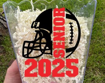 May include: Clear plastic container filled with shredded paper. The container has a football and helmet graphic in black and the word "SENIOR" in red. The year "2025" is also in red.