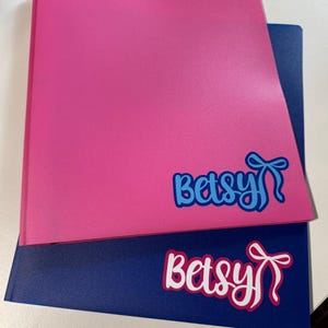 May include: Two folders, one pink and one navy blue, are stacked. Each folder has the name "Betsy" in a script font with a bow graphic. The pink folder has blue text, and the navy blue folder has white text.
