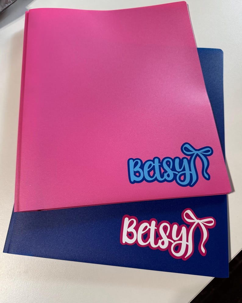 May include: Two folders, one pink and one navy blue, are stacked. Each folder has the name "Betsy" in a script font with a bow graphic. The pink folder has blue text, and the navy blue folder has white text.