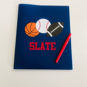 May include: A navy blue notebook featuring a basketball, baseball, and football graphic. The word "SLATE" is printed in red block letters below the sports icons. A red pen rests on the notebook.