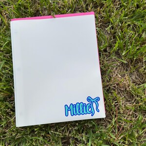 May include: A white binder with a pink spine rests on green grass. A blue and white sticker with the name "Millie" and a bow is on the lower right corner of the binder.