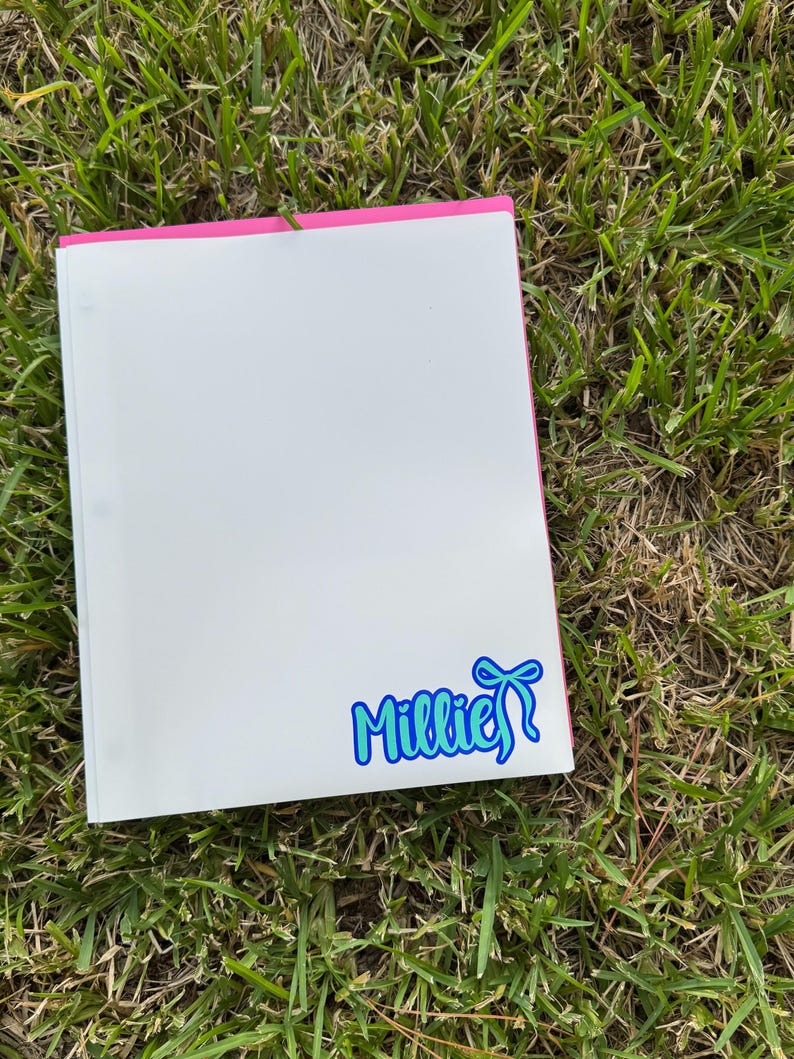May include: A white binder with a pink spine rests on green grass. A blue and white sticker with the name "Millie" and a bow is on the lower right corner of the binder.